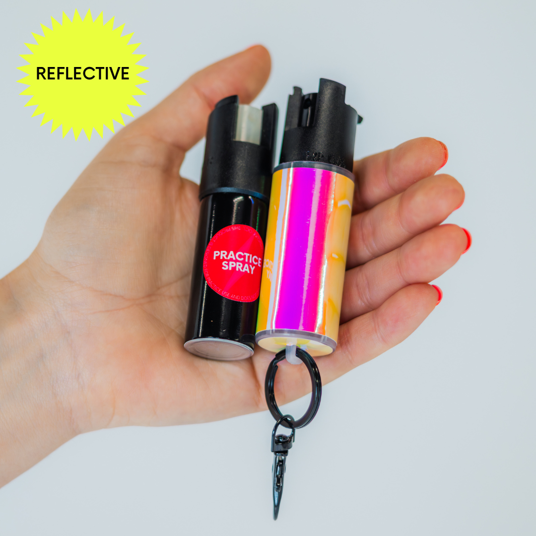 Hand holding a shiny yellow and pink pepper spray canister with a keychain with a practice spray canister with a reflective label on a white background