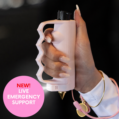 A hand holding a light pink personal safety alarm with a knuckle grip, a built-in flashlight, and a pink lanyard with a gold spike, set against a dark background with a pink 'NEW! LIVE EMERGENCY SUPPORT' badge.